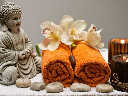 Eine Buddhafigur aus Stein, zwei gerollte Handtücher mit Orchideenblüten und Kerzen für die Ayurveda Gesundheitsberatung